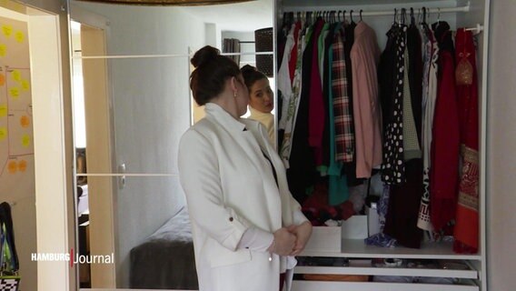 Eine Frau probiert vor ihrem Spiegel und dem offenen Kleiderschrank einen weißen Blazer an.  