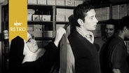 Nonne hilft einem jungen Mann in ein Jackett (in einer Kleiderkammer 1964)  