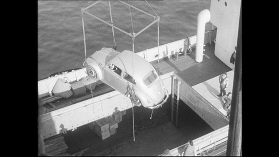Ein VW-Käfer hängt an einem Kran und wird auf ein Schiff geladen.  