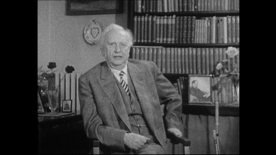 Karl Jaspers sitzt vor einem Bücherregal.  