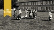 Frauen vor einem zweistöckigen Wohnheim, offenbar von der Gartenarbeit zurückkehrend (1964)  