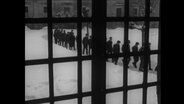 Blick durch ein Gitterfenster auf in einer Reihe laufende Männer.  