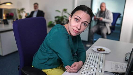 Eine Frau guckt fassungslos in die Kamera im Hintergrund ihre Bürokollegen.  