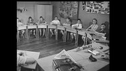 Schulkinder sitzen an Tischen in einem Halbkreis (Archiv-Bild).  