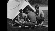 Drei Männer sitzen oberkörperfrei vor einem Zelt.  