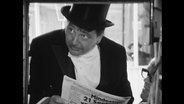 Wolfgang Neuss in Anzug und Zylinder liest eine Zeitung.  