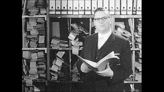 Ein Mann steht mit einem Buch in der Hand vor einem Regal voller Akten.  