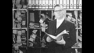 Ein Mann steht mit einem Buch in der Hand vor einem Regal voller Akten.  