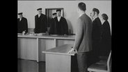 Richter und Prozess-beteiligte stehen in einem Gerichtssaal.  