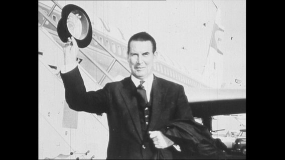 Gerhard Schröder steht vor einem Flugzeug und hebt seinen Hut.  