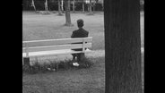 Aufnahme eines Mannes, der mit dem Rücken zur Kamera auf einer Bank sitzt.  