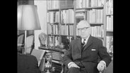 Professor Hallstein sitzt vor einem Bücherregal.  