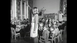 Eine Erzieherin mit weißer Schürze läutet eine Glocke vor Kindern, die an Tischen sitzen.  