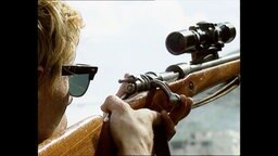 Ein Mann mit einer Sonnenbrille hält ein Gewehr in der Hand.  