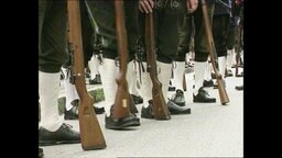 Gewehre stehen vor den Beinen von in Trachten gekleideten Männern.  