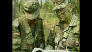 Ein mit Tarnfarben geschminkter US-Soldat und ein deutscher Soldat schauen gemeinsam in eine Karte  