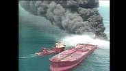 Ein Tanker auf de Meer, aus dem eine schwarze Rauchwolke aufsteigt.  
