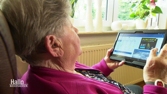 Eine Seniorin sitzt in einem Sessel und hat ein Tablet in der Hand.  