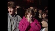 Eine Frau in pinker Jacke steht in einer Menschenmenge und hält sich die Hände vor ihr Gesicht (Artchivbild).  