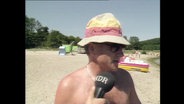 Ein Urlauber steht mit Sonnenhut am Strand und spricht in ein Mikrofon (Archivbild).  