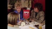 Drei Frauen, Mitglieder der SPD, sitzen am Tisch, auf dem eine SPD-Fahne steht (Archivbild).  