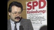 Der SPD-Ortsvereinsvorsitzende steht vor einem Plakat mit der Aufschrift "SPD Einladung" (Archivbild).  