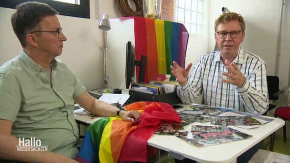 Zwei Männer sitzen an einem Tisch, auf dem Fotos und eine Regenbogenfahne liegen, im Hintergrund hängt eine weitere Regenbogenfahne.  