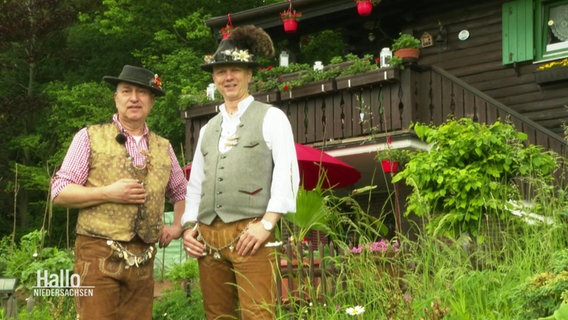 Zwei Männer in Tiroler Tracht stehen vor einer Holzhütte.  