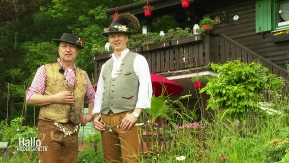 Zwei Männer in Tiroler Tracht stehen vor einer Holzhütte.  