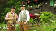 Zwei Männer in Tiroler Tracht stehen vor einer Holzhütte.  