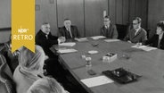 Acht Männer und eine Frau debattieren an einem großen Sitzungstisch (1964)  