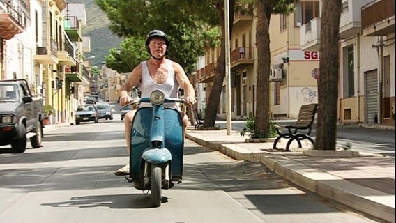 Ein älterer Mann im Unterhemd auf einer türkisblauen Vespa, erfährt durch eine italiniesche Stadt. © NDR 