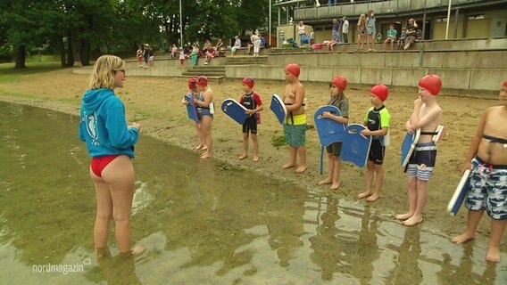 Kinder stehen in Badekleidung am Rande eines Sees.  