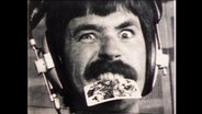 Ein Radiosprecher zieht eine Grimasse und hält eine Spielkarte zwischen seinen Zähnen fest  