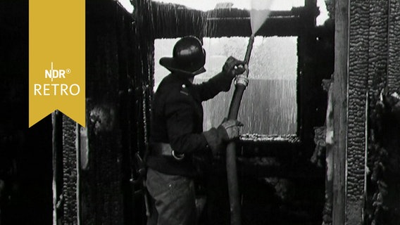 Feuerwehrmann spritzt nach oben in einem verkohlten Gebälk (1963)  