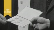 Briefmarkenheft wird in einer Hand gehalten, eine andere Hand zeigt mit Stift auf eine Marke (1963)  