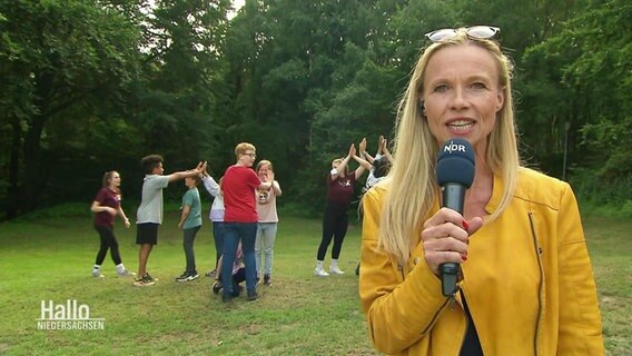 Eine Reporterin berichtet live, im Hintergrund spielen mehrere Jugendliche ein Laufspiel.  
