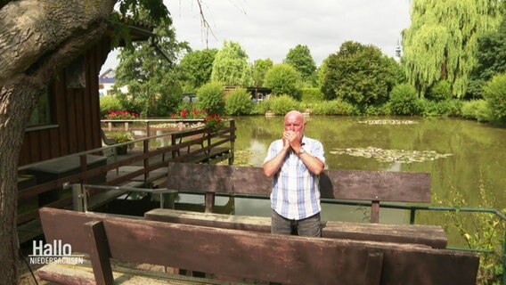 Georg Malkowsky steht an einem großen Teich, umgeben von grüner Natur. Er steht auf einem Steg und spielt die Mundharmonika.  
