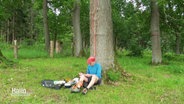 Rudolf Rantzau sitzt am Fuß eines Baumes und macht sich Notizen. Vom Baum herunter hängt ein rotes Kletterseil.  