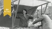 Marktfrau in einem Stand, in den der Sturm pfeift, ruft aufgeregt, ein Kunde versucht noch, die Auslage vor dem Windstoß festzuhalten (1958)  