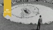 Besucher sitzen im Kreis um eine runde Fläche, die ein Steinzeitgrab darstellt (1963)  