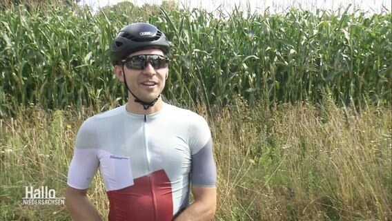 Eike Lindemann gibt ein Interview. Er trägt einen Fahrradhelm, Sonnenbrille und ein komplettes Sportoutfit für lange Radtouren.  