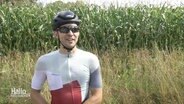 Eike Lindemann gibt ein Interview. Er trägt einen Fahrradhelm, Sonnenbrille und ein komplettes Sportoutfit für lange Radtouren.  