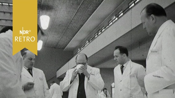 Mehrere Männer in weißen Kitteln stehen um einen Tisch bei einer Lebensmittelverkostung (1964)  