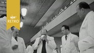 Mehrere Männer in weißen Kitteln stehen um einen Tisch bei einer Lebensmittelverkostung (1964)  