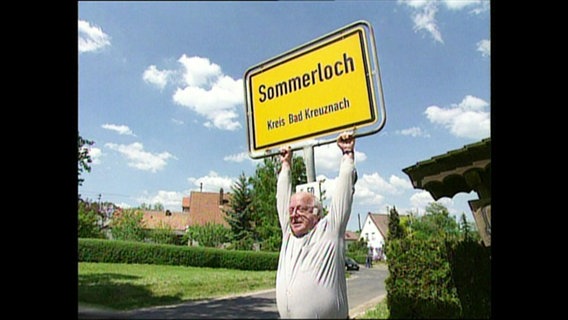 Ein Mann hängt an dem Ortsschild "Sommerloch"  