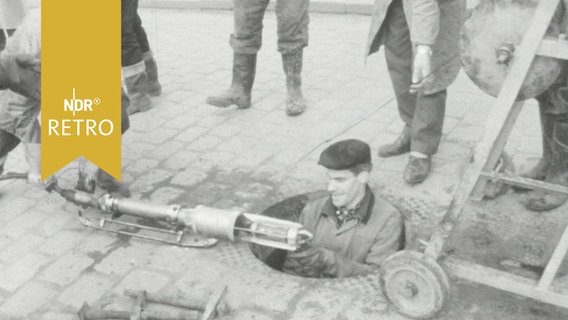 Ein Kanalarbeiter steigt inmitten von Kollegen in einen Sielschacht ab (1964)  