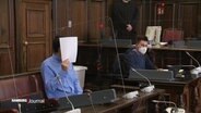 Ein Mann sitzt in einem Gerichtssaal und hält sich ein weißes Papier vor das Gesicht.  