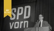 Gerbert Wehner vor einem Plakat "SPD vorn" bei einer Rede auf dem Parteitag des neuen SPD-Bezirks Nordniedersachsen 1964  