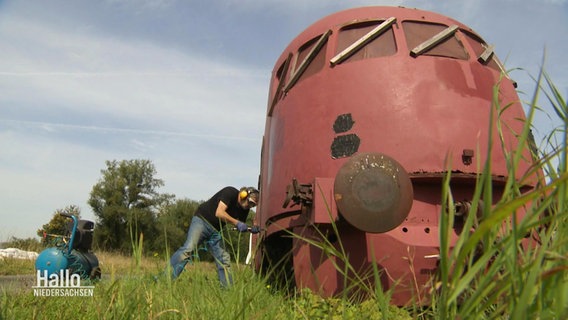 Ein Mann arbeitet an einem alten Zug, dessen Oberfläche komplett abgeschliffen wurde und wo die Fenster herausgebaut wurden.  